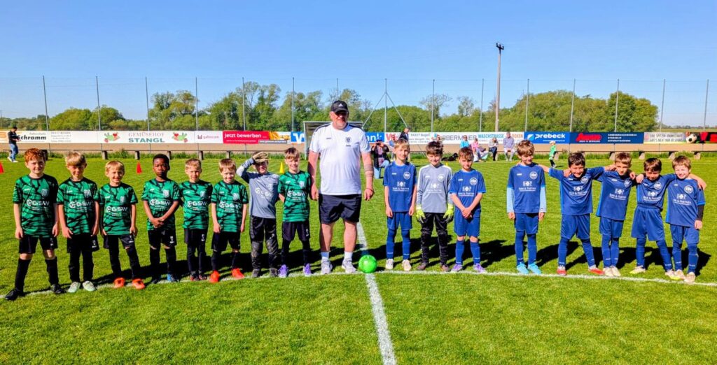 Mannschaftsfoto der DJK Straubing U9 gemeinsam mit dem Trainerteam und dem SC Kirchroth nach dem Spiel auf dem Sportplatz in Kirchroth, im Hintergrund die markante Pfarrkirche St. Margaretha bei strahlendem Frühlingswetter.