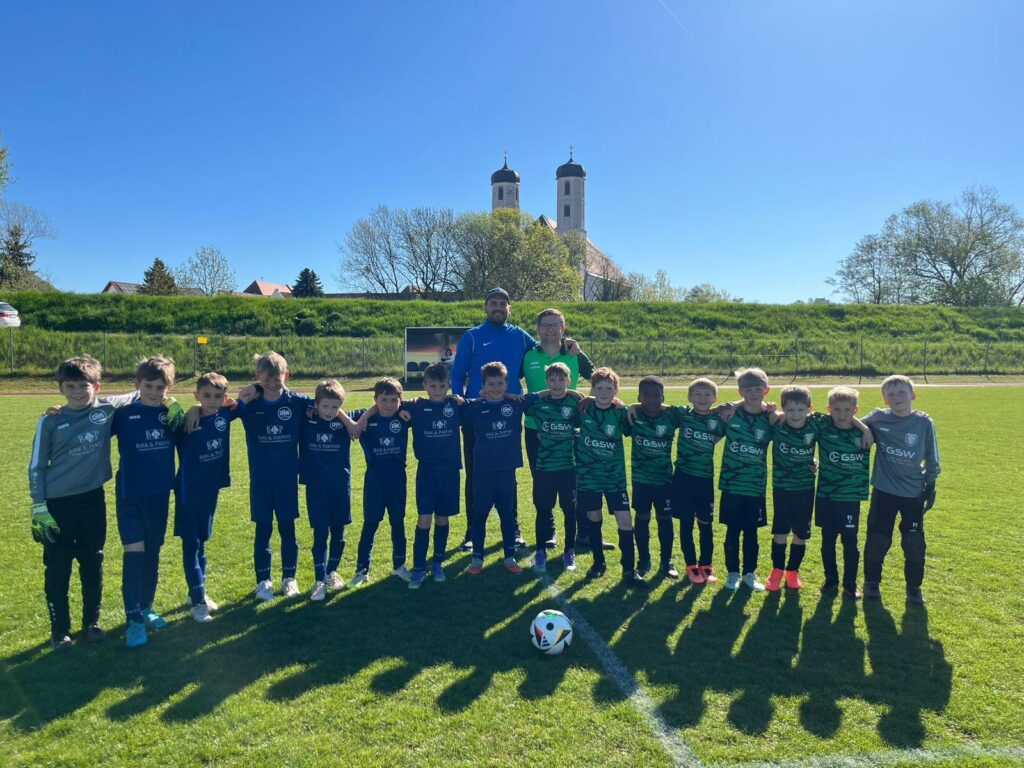 Aufstellung beider Mannschaften vor dem Anpfiff beim Auswärtsspiel der DJK Straubing U9 in Bogen – die Kinder in grün-schwarzen und blauen Trikots posieren gemeinsam mit dem Schiedsrichter auf dem Hauptspielfeld, im Hintergrund die Werbebanden der Sponsoren.
