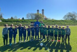 Aufstellung beider Mannschaften vor dem Anpfiff beim Auswärtsspiel der DJK Straubing U9 in Bogen – die Kinder in grün-schwarzen und blauen Trikots posieren gemeinsam mit dem Schiedsrichter auf dem Hauptspielfeld, im Hintergrund die Werbebanden der Sponsoren.