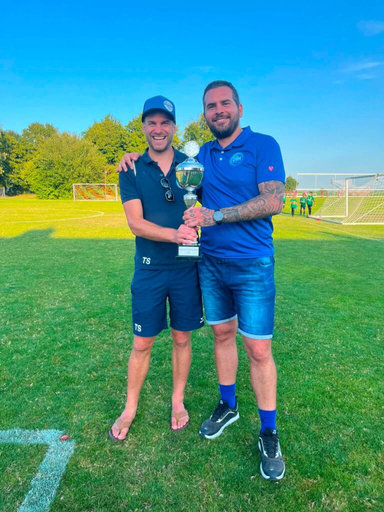 Die Trainer Tom Straubinger und Sinisa Stojanovic der DJK SB Straubing F1 halten stolz den Siegerpokal des Kiwanis Cup Alburg 2026 in die Kamera.