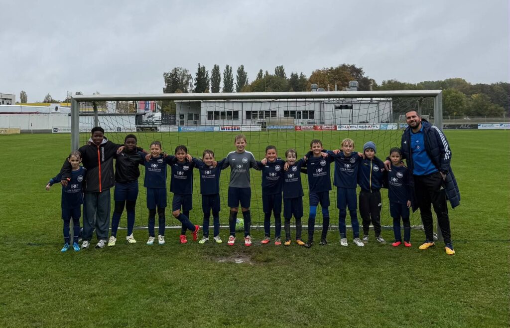 U11-Mannschaft (E-Jugend) der DJK SB Straubing in dunkelblauen Trikots Arm in Arm vor dem Tor, mit Trainer, an einem regnerischen Herbsttag